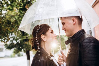 Genç mutlu çift şehir caddesinde şeffaf bir şemsiye altında öpüşüyor. Erkek ve kadının yakın plan portresi. Şemsiyenin altında gülümseyen sevimli bir çift. Şehirde güneşli bir sonbahar günü.