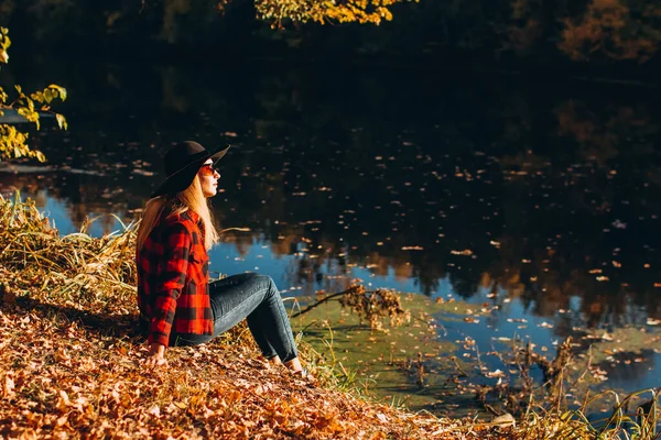 Gri şapkalı bir sarışın sonbahar ormanında yürüyor. Kırmızı ceketli neşeli kız güneşli bir sonbahar gününde gölde oturur. Ormanda yürüyen bir kadın.