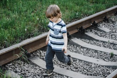 curly-haired boy in a striped T-shirt walks on rails. railway, danger