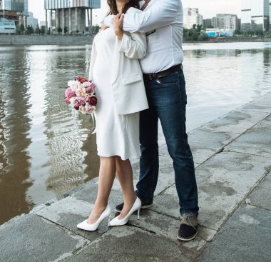 İnce, beyaz bir elbiseyle, ince spagetti askılarıyla, omuzları çıplak ve şakayık demeti olan hamile gelin. 