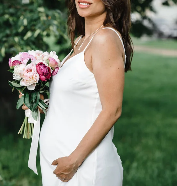 Pregnant bride in a white thin dress on thin straps with bare shoulders with a bouquet of peonies. motherhood — Stock Photo, Image