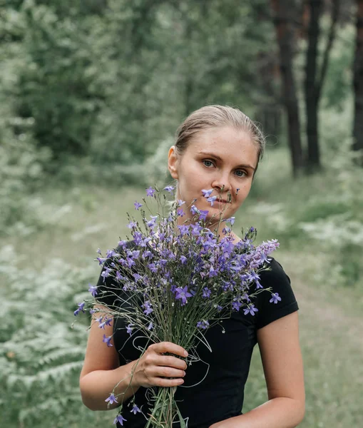 Siyah tişörtlü, kuyruklu genç bir kadın ve ormanda elinde mavi kır çiçekleri tutan gri bir kot. — Stok fotoğraf
