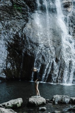 Dağ şelalesinin arka planında at kuyruklu genç bir kadın. 
