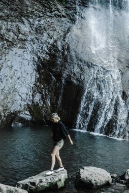 Dağ şelalesinin arka planında at kuyruklu genç bir kadın. 