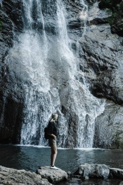 Dağ şelalesinin arka planında at kuyruklu genç bir kadın. 