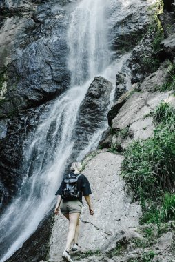 Dağ şelalesinin arka planında at kuyruklu genç bir kadın. 