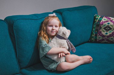 Küçük tatlı kızıl saçlı bir kız kanepede oturuyor ve bir ayıya sarılıyor.