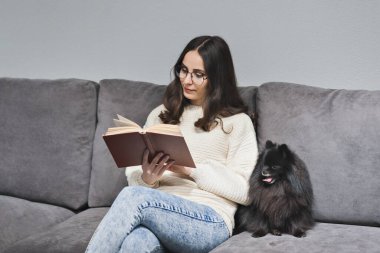 Kız kanepede bir köpeğin yanında kitap okuyor.