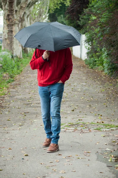 Man het dragen van een rode Pullover en blauwe jeans wandelen in de straat met een zwarte paraplu — Stockfoto