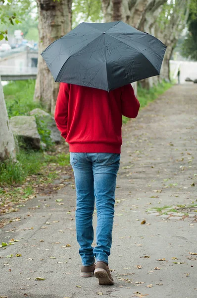Man het dragen van een rode Pullover en blauwe jeans wandelen in de straat met een zwarte paraplu — Stockfoto