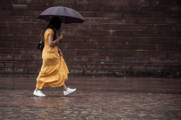 Женщина в жёлтом платье гуляет с зонтиком на булыжнике в городе — стоковое фото