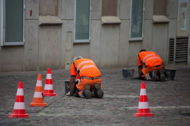 Arka planda, sokakta eski kaldırım taşları döşeyen işçilerin portresi.