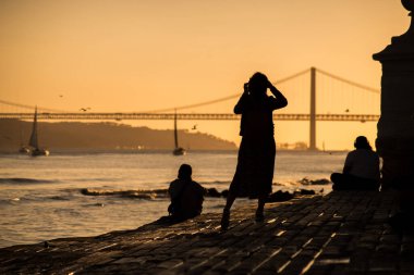 Lizbon - Portekiz - 30 Eylül 2020 - Günbatımı arkaplanındaki ünlü 25 Nisan süspansiyon köprüsünün akıllı telefonuyla fotoğraf çeken turist silueti