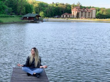 Yalnız kız bir gölün kenarında meditasyon yapar ve nilüfer pozisyonunda açık gözlerle gülümser..
