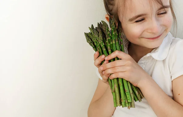 Gülümseyen küçük bir kızın ellerinde yeşil kuşkonmaz. Bir sürü taze kuşkonmaz. Sağlıklı organik yiyecekler. Evde yemek pişirmek. Doğal vitaminler, yemek için ham malzeme. Özenle seçilmiş biyolojik kuşkonmaz..