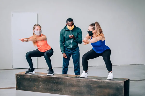 Two girls exercising with a personal trainer at the gym with protective mask in pandemic period of covid19 - Two fit young girls in a gym doing squats with their afro american trainer