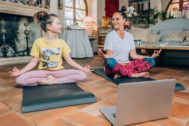 Anne ve kızı koronavirüs salgını sırasında karantina sırasında evde yoga dersleri alıyor. Aile izolasyon sırasında internetten spor yapıyor. Güzel genç kadın ve kızı evde yoga yaparken gülümsüyorlar.