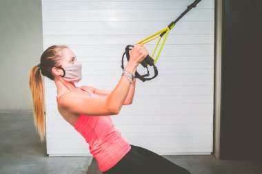 Kollarını koruyucu maskeyle çalıştıran ve salgın hastalık salgını sırasında spor salonunda Trx Fitness 'ı bağlayan bir kız 19 yaşında şınav çeken bir kadın göğüs üst göğüs kasları kaslarını çalıştırıyor.