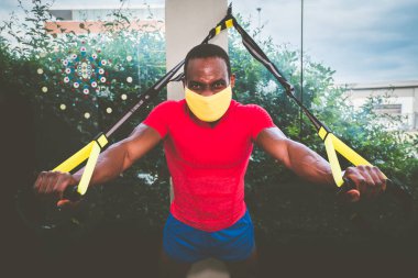 Afrika kökenli Amerikalı bir adam kollarını koruyucu maskeyle eğitiyor ve karantina sırasında spor salonunda Trx Fitness kemerlerini bağlıyor. 19 numaralı salgın için.