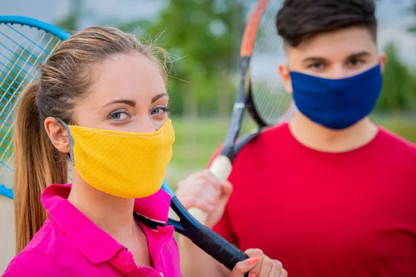 Tenis oyuncularının koruyucu maskelerle dışarıda raket tutarken portresi - karantina sırasında tenis oynayan sarışın kız ve genç oğlanı yakından takip etmek, açık havada tenis oynamaya hazırlanmak - virüs salgını