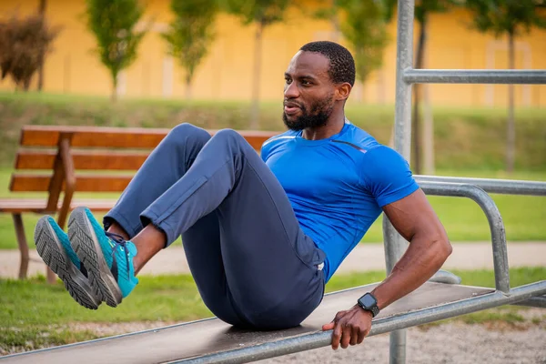 Fitness afro american boy doing situps outdoor - Handsome african man doing stretching exercises at open air gym in the park. Abdomen Exercises on the bench, training crunch in the City Park