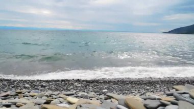 Denizin küçük dalgaları bir çakıl taşı plajına su sıçratır. Uzaktan görünen dağlar