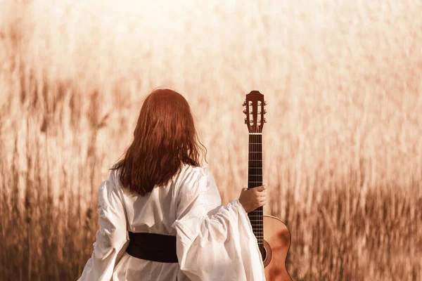 白い中世のドレスに赤い髪をした美しい若い女性がギターを持ち 日没の暖かい光の中で日当たりの良いフィールドを歩いています レンズフレア トーン画像 選択的フォーカス ぼやけたボケ — ストック写真
