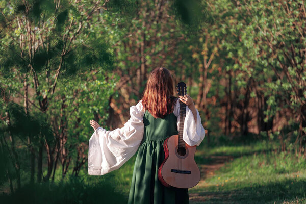Young woman in medieval long dress walks with guitar in sunny forest. Fantasy girl or bard at summer nature. Back view.