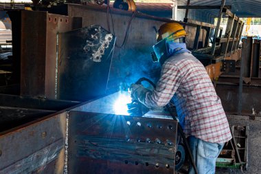 Kaynakçı, Flux Cored Arc Welding (FCAW) işlemiyle bir çelik yapı çalışması yapıyor ve endüstriyel fabrikada güvenlik için kişisel koruyucu ekipman (PPE) ile uygun şekilde giyiniyor.