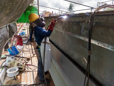 Kaynakçı, Flux Cored Arc Welding (FCAW) işlemiyle bir çelik yapı çalışması yapıyor ve endüstriyel fabrikada güvenlik için kişisel koruyucu ekipman (PPE) ile uygun şekilde giyiniyor.