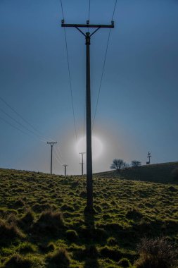 Elektrik kabloları ve yalıtımlı ahşap elektrik direğinin arkasındaki güneş. Arka planda mavi gökyüzü.