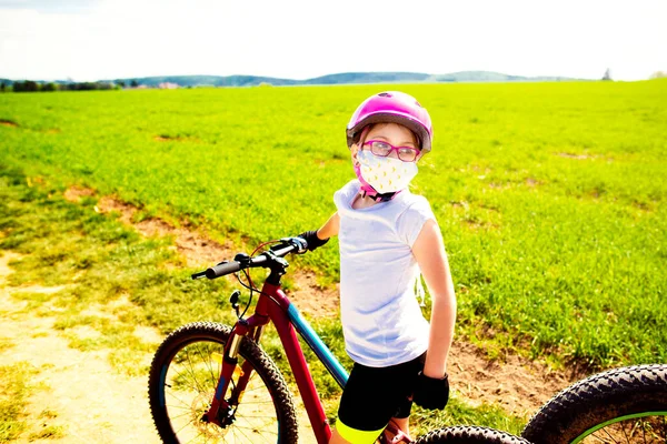 Sport and quarantine. 9 years biker girl with mask amidst of the fields in the sunny day. Freedom, recreation and sport concept.