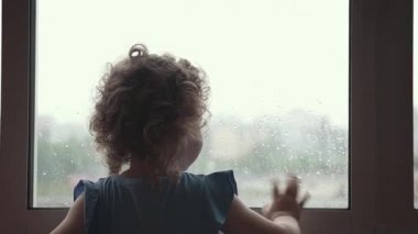 Yağmurlu bir günde pencereden dışarı bakan küçük bir kızın silueti. Pencerenin yanında yalnız bir çocuk duruyor ve elini sallayarak birine veda ediyor..