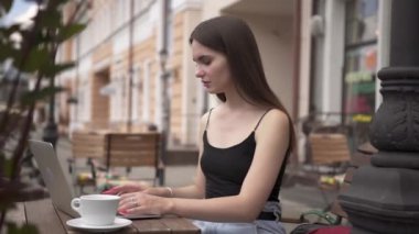 Açık kafede dizüstü bilgisayar kullanan çekici bir kız öğrenci. Genç kız kafede dizüstü bilgisayarla çalışıyor. Modern teknoloji ve gençlik kavramı.