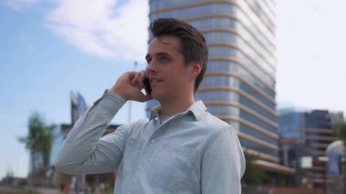 Yakışıklı genç adamın yavaş çekim görüntüsü modern bir şehrin arka planına karşı telefonda konuşuyor. Komik bir şey duyar ve güler..