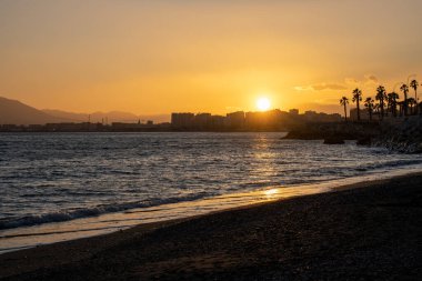 Yaz batımı. Denizin üzerinde turuncu gökyüzü, ufukta palmiye ağaçları, şehir.