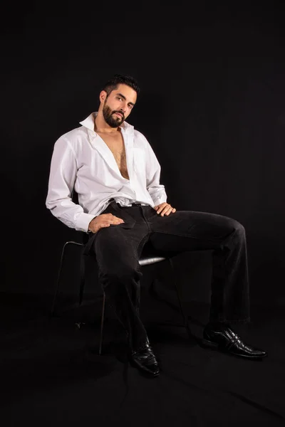 Handsome seductive man with open shirt showing chest sitting on a chair. Attractive stylish bearded man in formal wear. Black background.