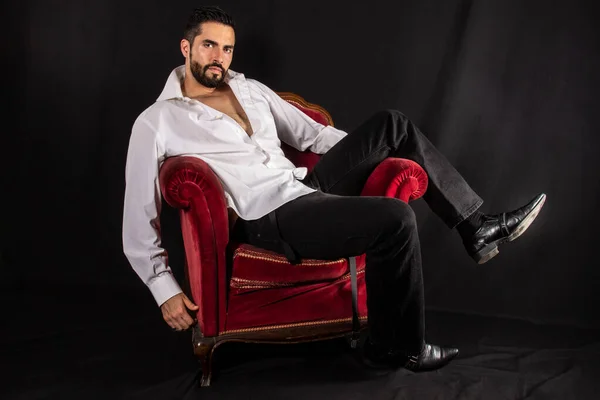 Handsome seductive man with open shirt showing chest sitting on a luxury red armchair. Attractive stylish bearded man in formal wear. Black background.