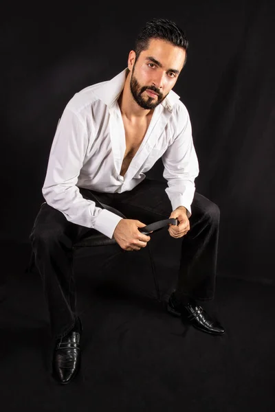Handsome seductive dominant man with open shirt showing chest sitting on a chair. Attractive stylish bearded man in formal wear. Black background.