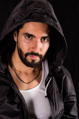 Siyah kapüşonlu yakışıklı sakallı adamın dramatik portresi siyah arka planda kameraya bakıyor.