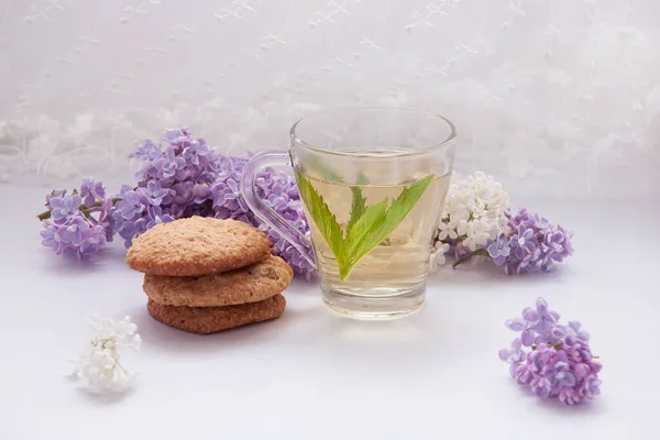 Şeffaf fincanda naneli çay ve arka planda yulaf ezmeli kurabiye. Leylak çiçeklerinin etrafında.