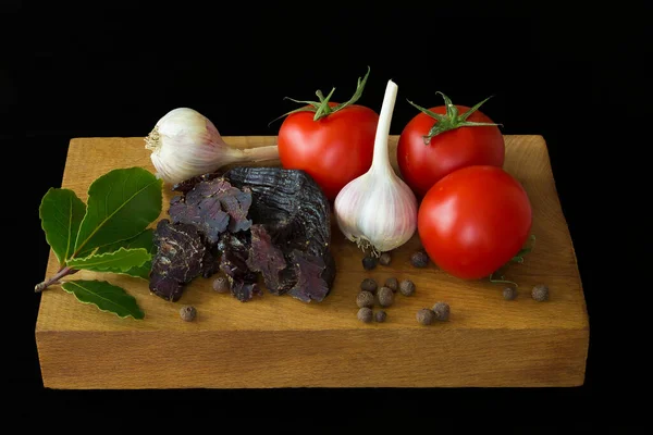 Domates, sarımsak, kurutulmuş geyik eti, körfez yaprakları ve ahşap bir tahtanın üzerindeki tüm baharatlar. Siyah arka plan. İzole et. yan görünüm.
