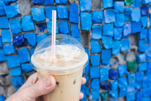 Holding iced coffee in takeaway cup on wall background with colored mosaics