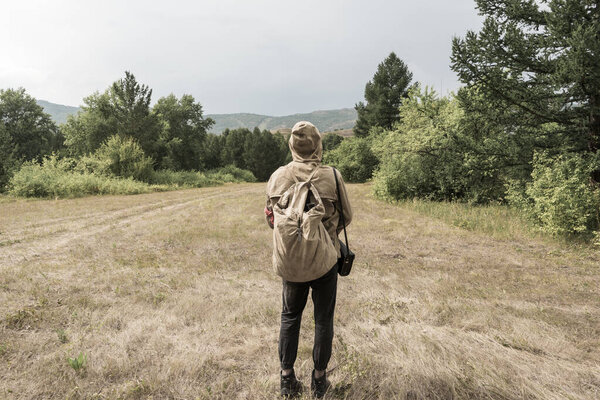 Back view of tourist man with a backpack standing ready to travel on green forest background. Stalker in the post-apocalyptic world.