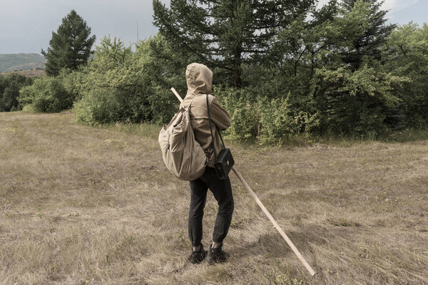 Back view of tourist man with a backpack standing ready to travel on green forest background. Stalker in the post-apocalyptic world.