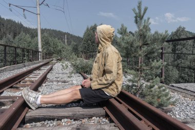 Adam terk edilmiş bir demiryolu köprüsünde oturuyor. Yolun karşısındaki ormanda eski bir demir köprü..