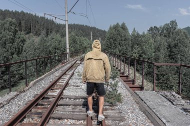 Bir adam terk edilmiş bir demiryolu köprüsünden geçiyor. Yolun karşısındaki ormanda eski bir demir köprü..