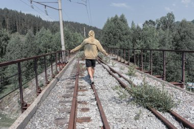 Bir adam terk edilmiş bir demiryolu köprüsünden geçiyor. Yolun karşısındaki ormanda eski bir demir köprü..