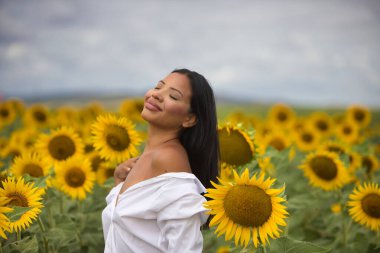 Koyu tenli, beyaz gömlekli ve çıplak omuzlu güzel bir Latin kadın güneşli bir günde ayçiçeklerinin arasında duruyor. Şehvetli kadın gülümser ve mutlu görünür.