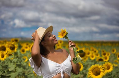 Beyaz gömlek ve hasır şapka giyip ayçiçeklerinin arasında duran koyu tenli genç bir Latin kadın çiftçinin portresi. Kız elinde bir çiçek tutuyor ve kokluyor ve ona bakıyor.